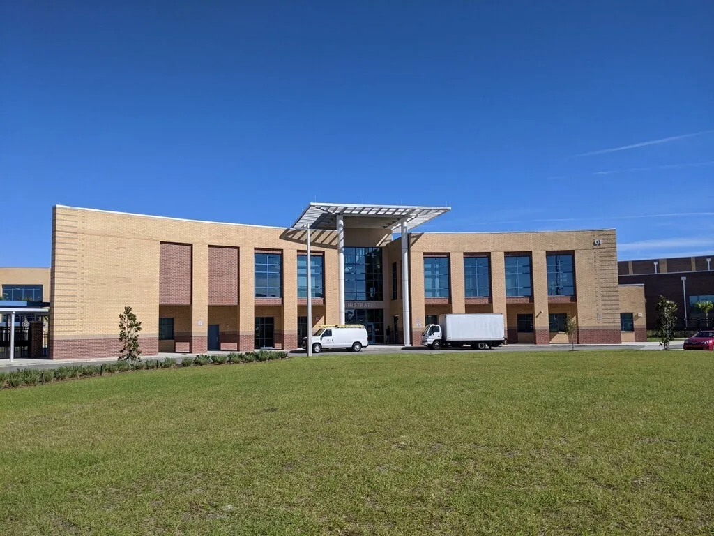Brown brick school building surrounded by blue sky and green grass with white van and white truck parked in front