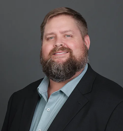 Man with brown hair, beard and moustache wearing black jacket and blue shirt with dark gray background