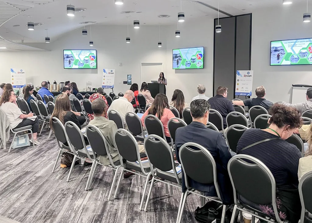 Amanda Polematidis speaking at the front of a conference room with two rows of people sitting in chairs and several monitors on wall behind her