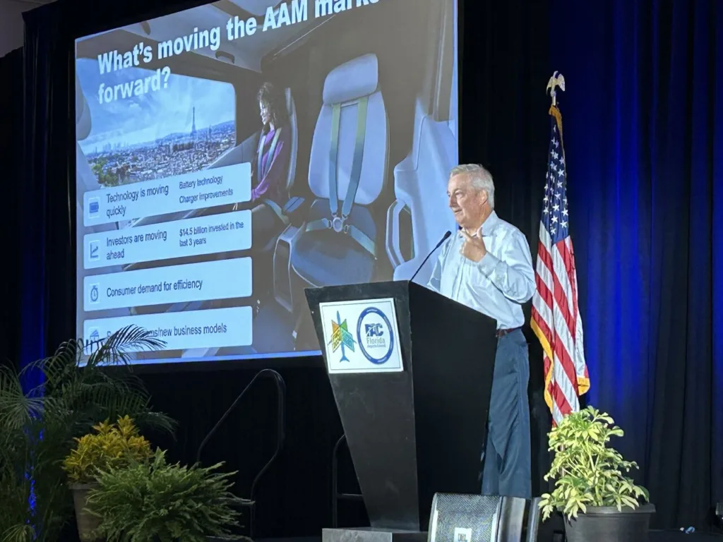 Man behind lecturn with screen behind him that says. "What's moving the AI machine forward?"