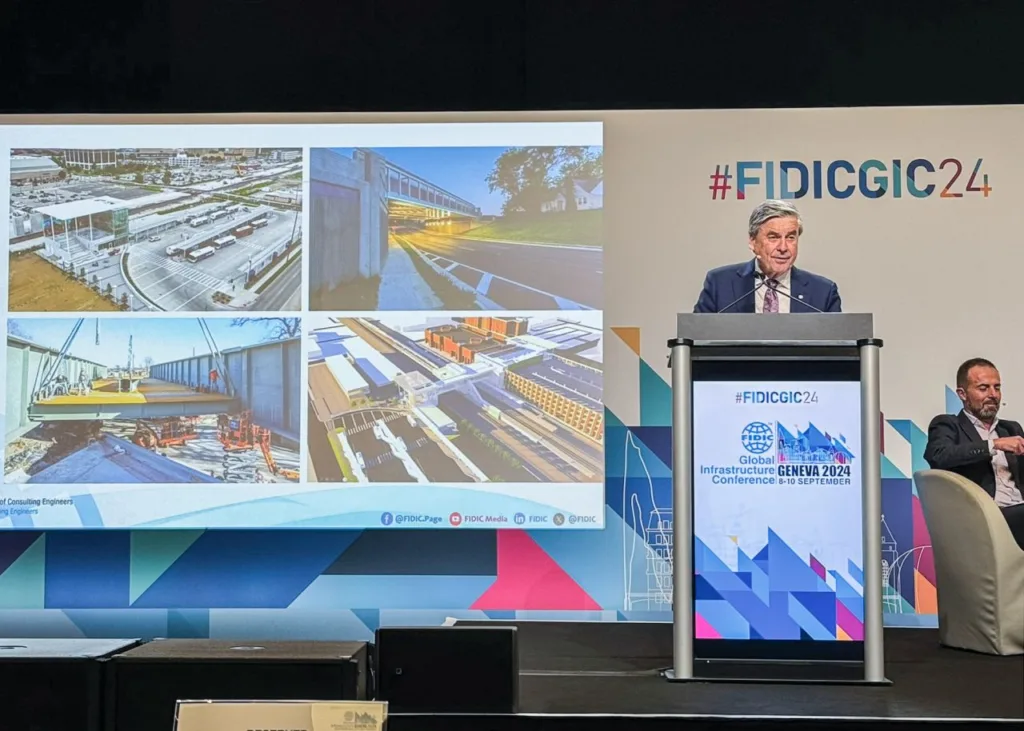 Man speaking from behind a lecturn with an image with FIDICGIC24 Global Infrastructure Conference on the front, with screen off to the left showing images of project work.