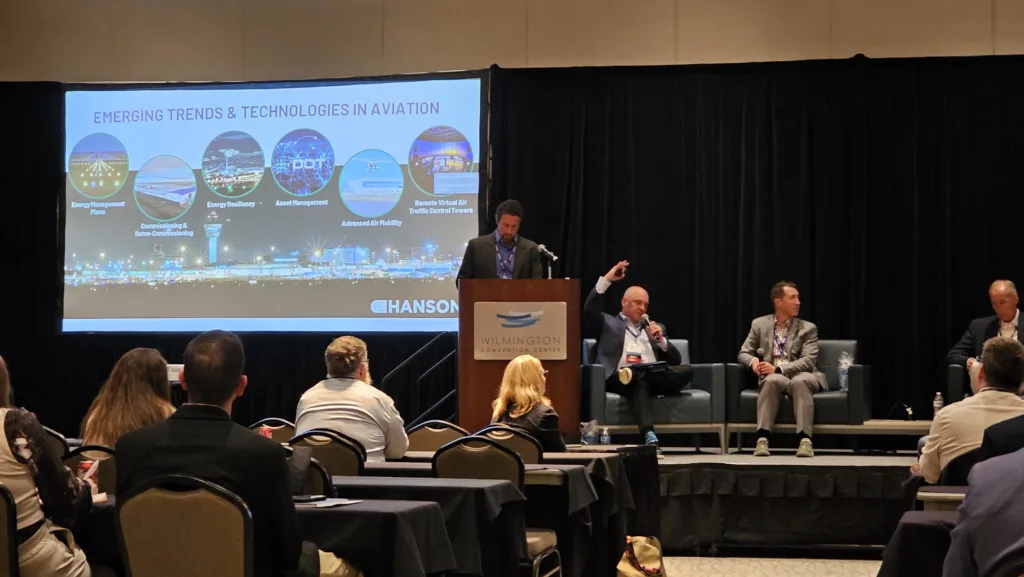 Four men on stage, one standing behind a lecturn and three sitting, discussing topic on screen behind them, "Emerging trends and technologies in aviation." Several attendees sitting at tables in front of stage.