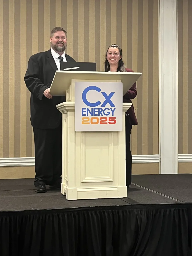 Man and woman standing behind lecturn with sign on front that says, "Cx Energy 2025" and brown and tan striped wallpaper in the background