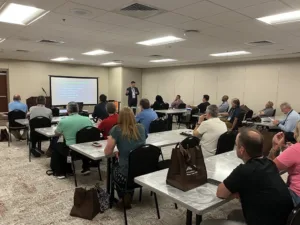 Kurt Bialobreski speaking in front of conference room to several rows of attendees sitting at tables, with monitor in background