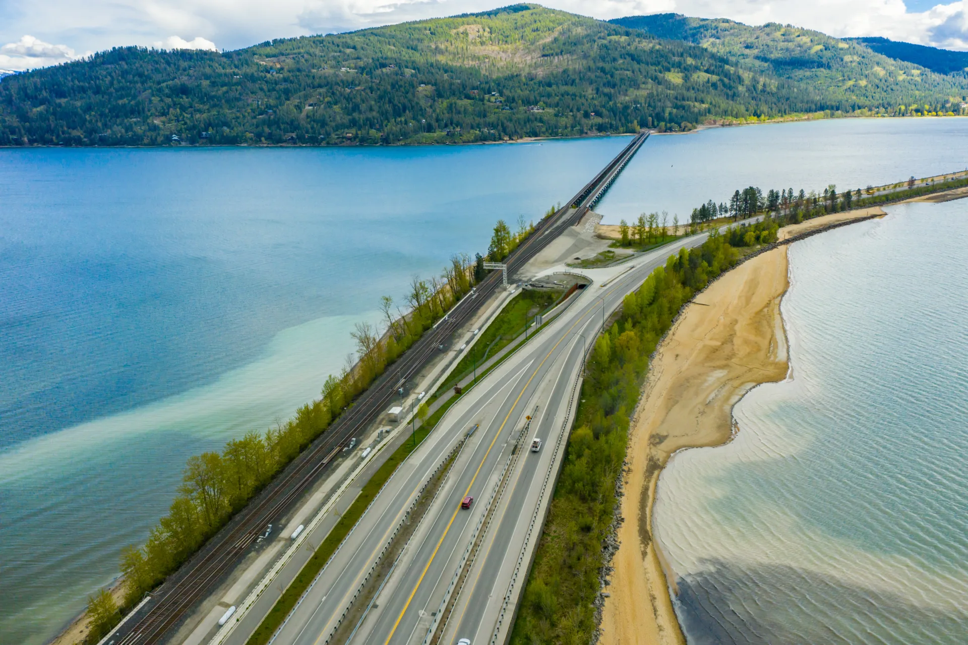 Aerial photo of cars crossing bridge over water beside train tracks with grassy hills in background