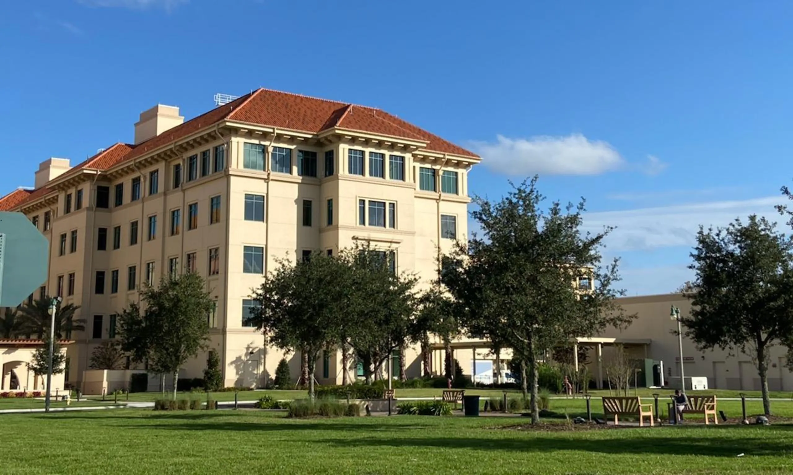 Exterior of medical facility-beige with orangish-red roof and benches on grass in foreground. Blue sky with clouds in background