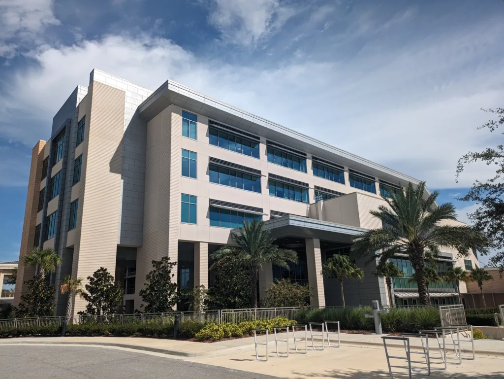 Hospital building exterior - tan with many glass windows, surrounded by palm trees and blue sky with clouds