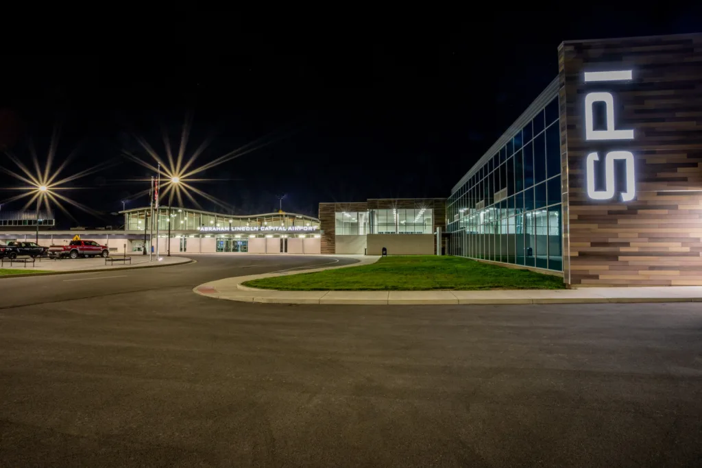 Airport at night with "SPI" in large, lit-up letters on side of building in foreground and parking lot lights glowing in background