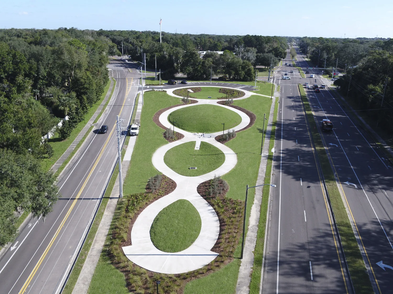 Aerial view of grassy park with curving sidewalks and newly planted trees, lined on each side with a roadway