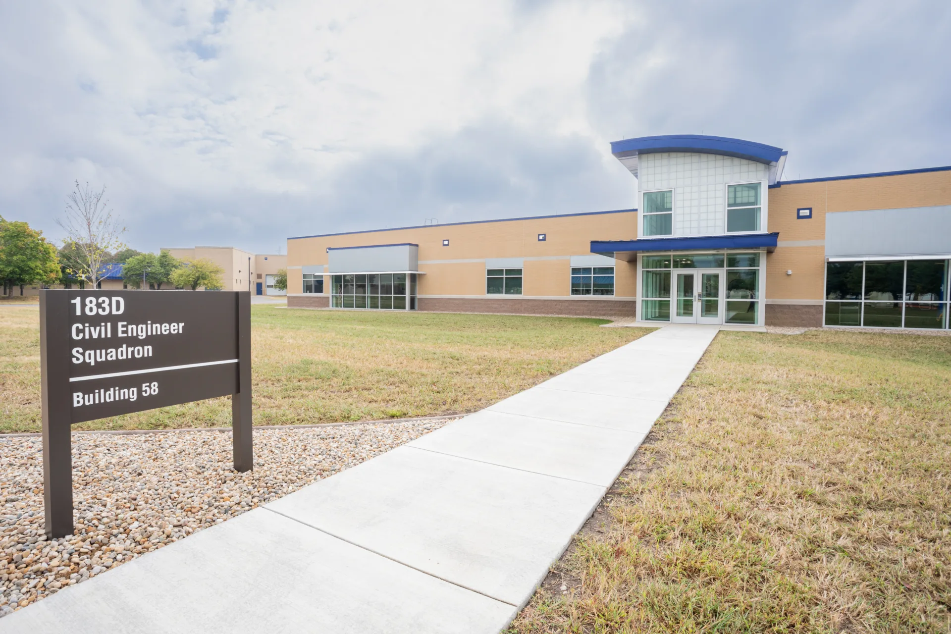 Exterior view of building with tan and brown brick, windows and tall entrance with royal blue trim. Sign in front, next to sidewalk leading to entrance, reads, "I83d Civil Engineer Squadron Building 58."