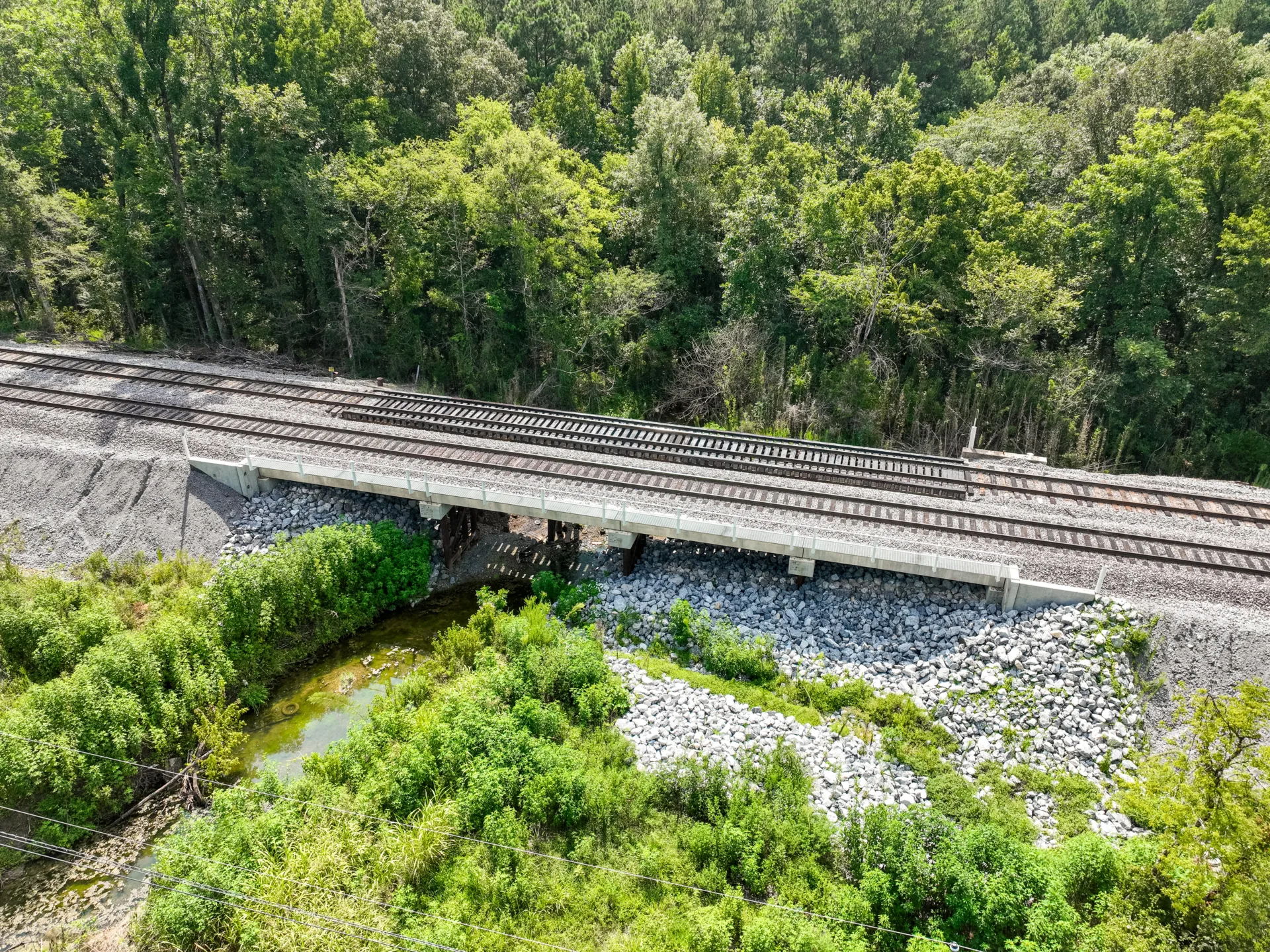 Aerial view of railroad bridge with trees in background and shrubs and rocks in foreground