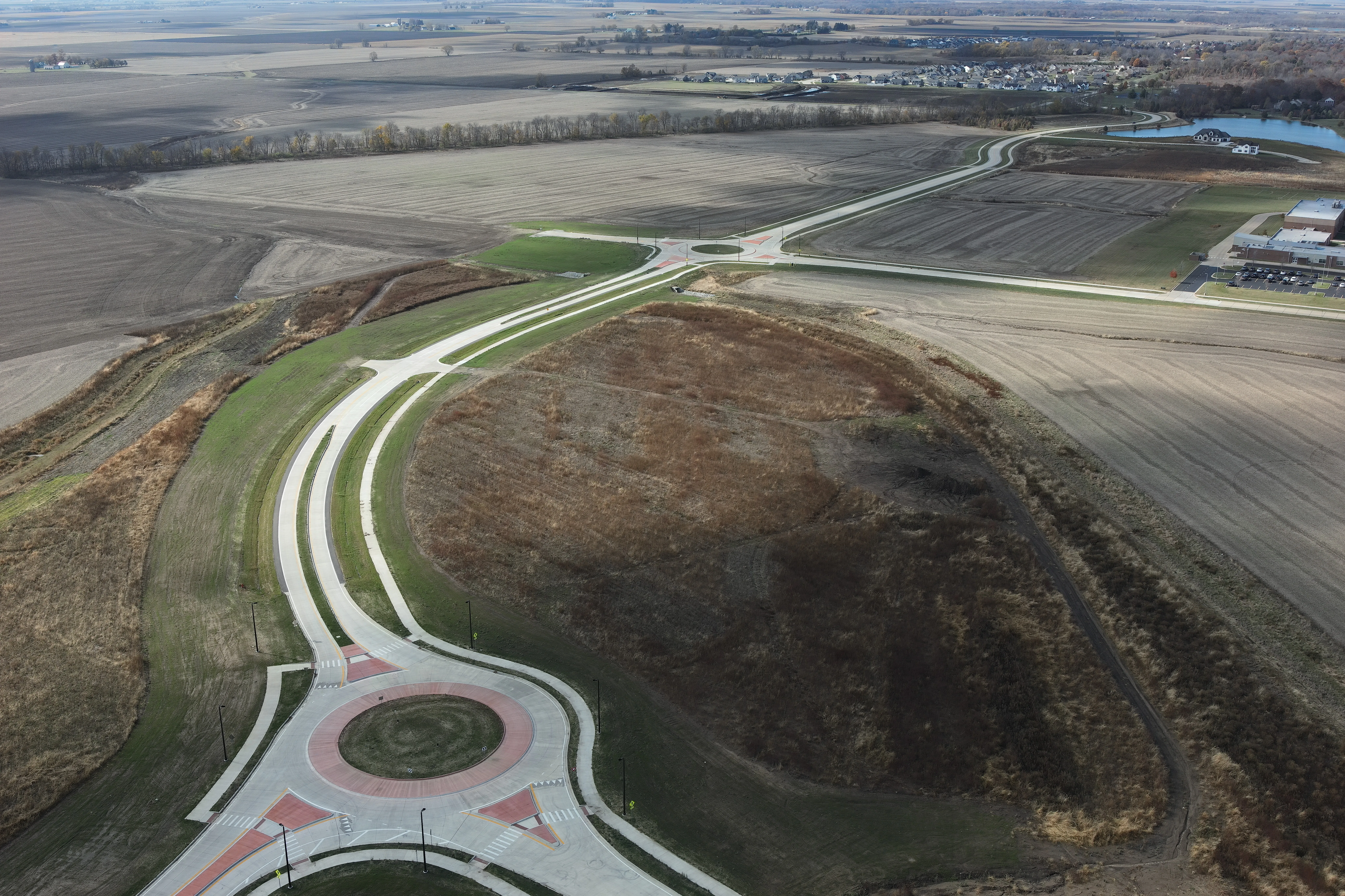 Aerial view of a new road with one roundabout intersection in the foreground and a second in the distance. Farm fields surround the roadway, and buildings and a pond are farther in the distance.