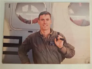 Close-up photo of Dennis Hollahan standing in front of a military plane wearing uniform and holding an item with a strap over his shoulder