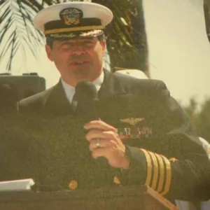 Eric Menger holds microphone at lectern outdoors while wearing military uniform and hat with palm trees in background