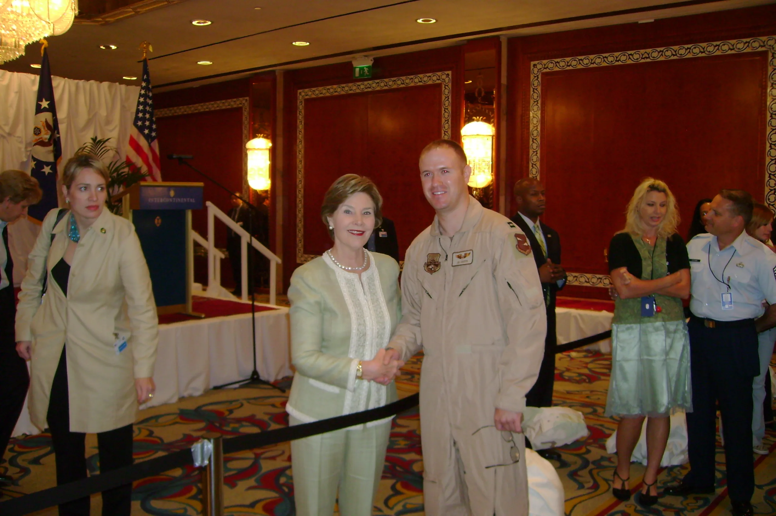 Joe Crispen in tan military jumpsuit inside wood-paneled conference room, shaking hands with Laura Bush, wearing light green pant suit with several people in background