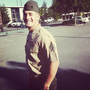 Outdoor photo of Kyle Geitner in brown military uniform with building, parking lot and cars in background