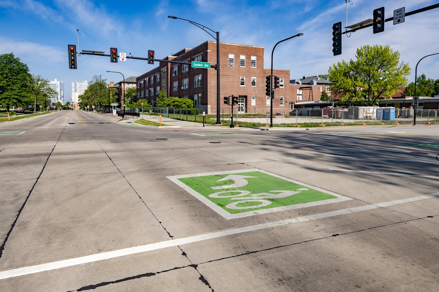 Campus intersection with green and white bicyclist graphic painted on concrete and traffic lights at left and right.