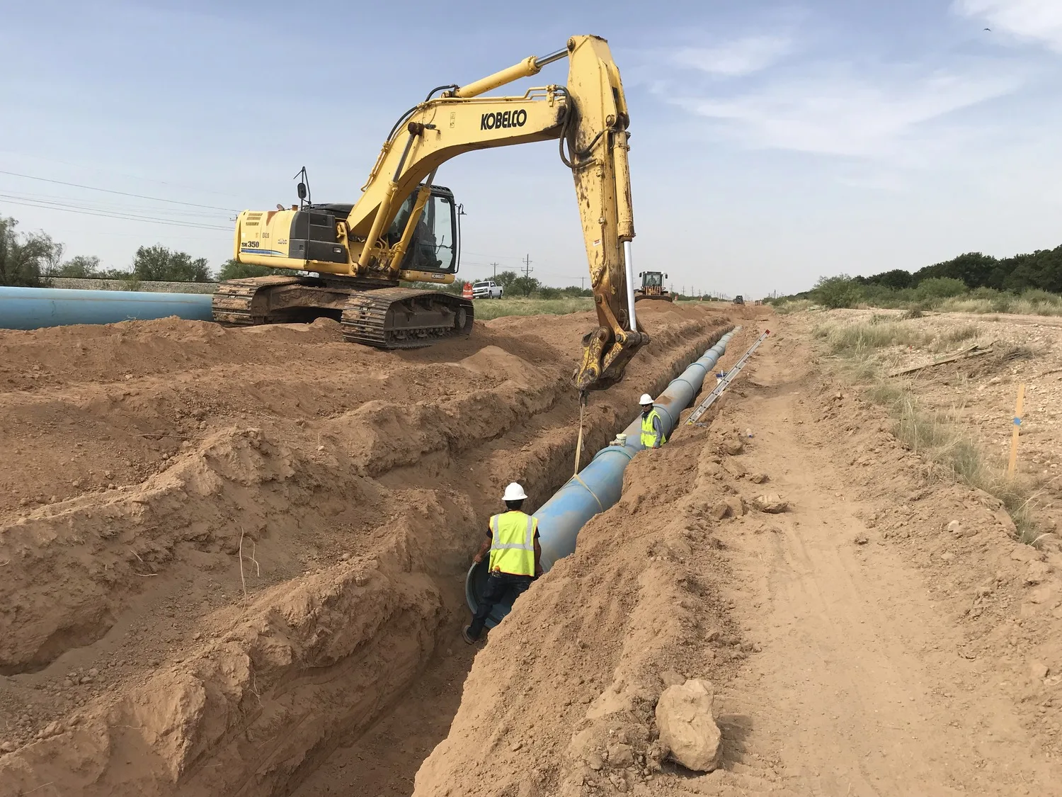 Construction site with large dirt trench and workers with blue pipe in trench; construction machine maneuvering pipe