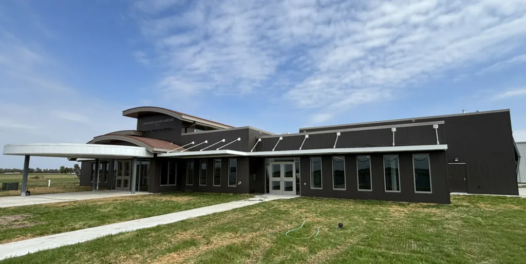 View of front of brand-new airport terminal building with dark exterior and vertical windows lining the front; green grass in the foreground and blue sky with puffy white clouds in the background