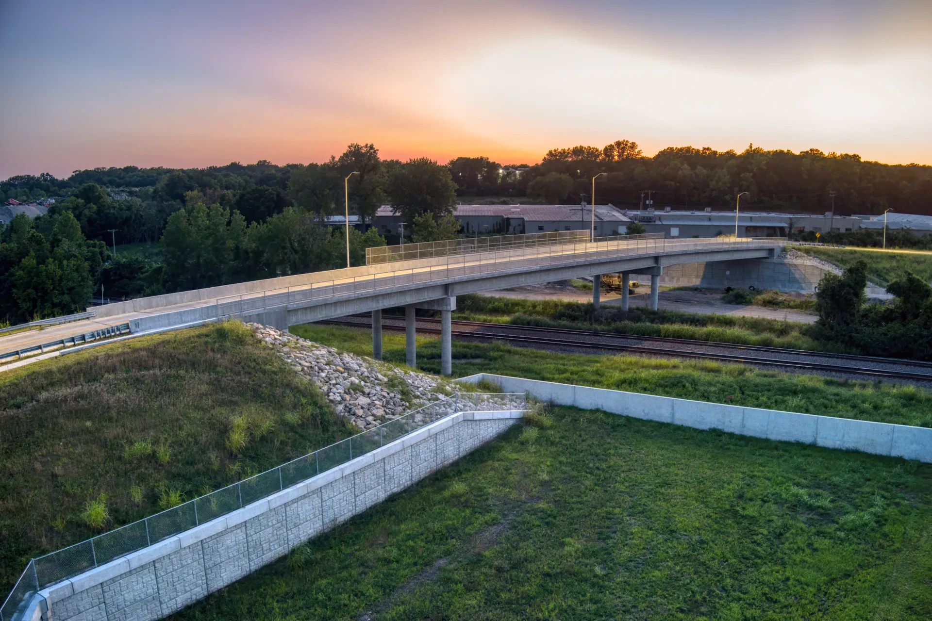 Sunset photo of bridge with railroad tracks running underneath and green grass with retaining wall in foreground and trees in background