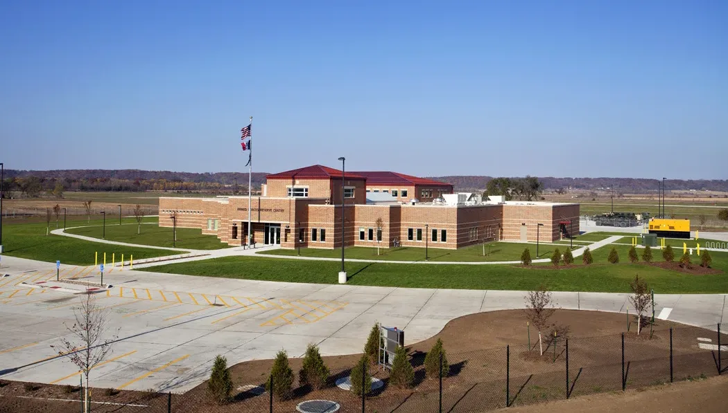 Aerial view of the Muscatine Iowa joint readiness center, with sprawling red brick building and blue sky in background; flagpole with three flags in foreground and parking lot at bottom