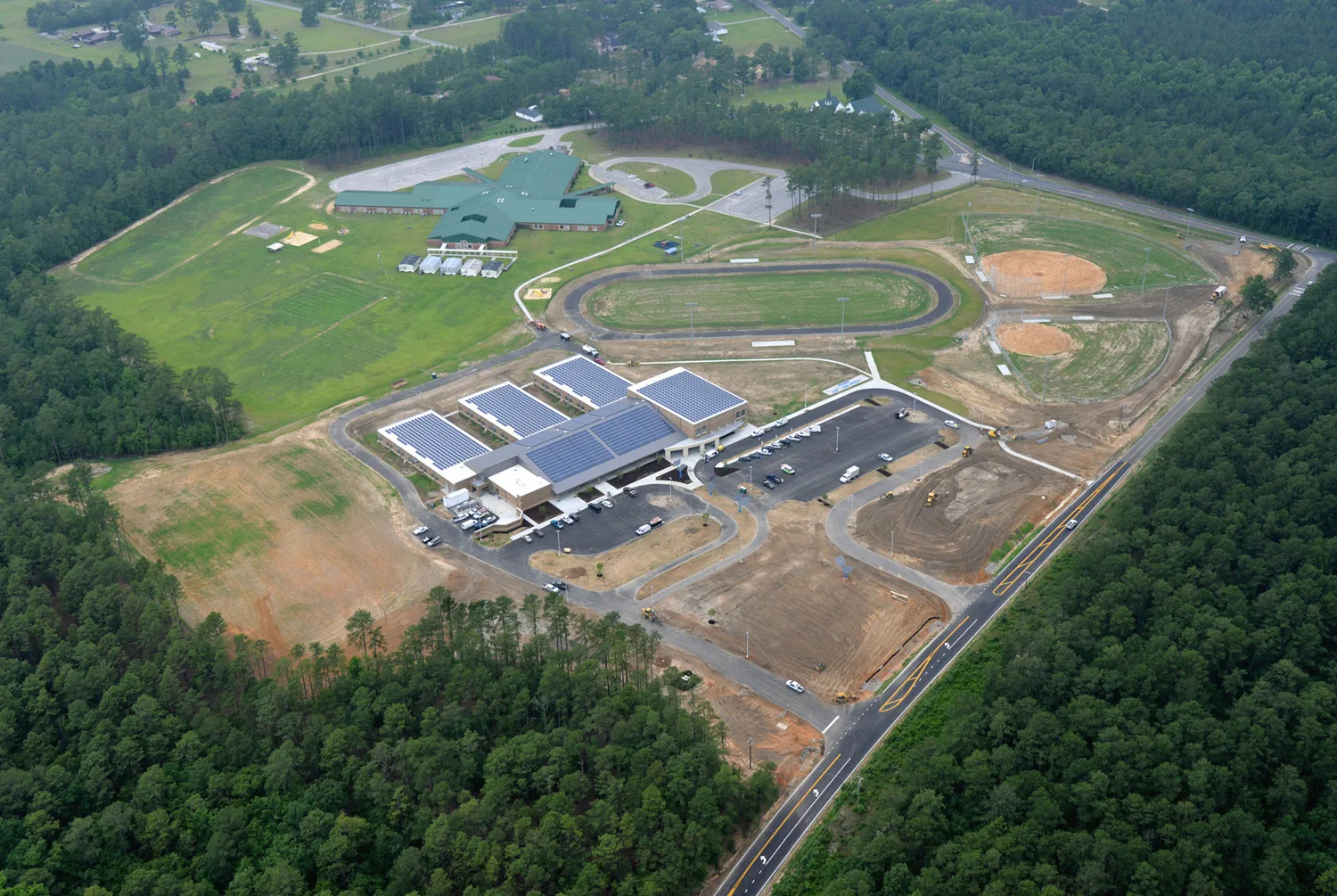 Aerial view of a building with solar panels covering roof; trees on all sides and track toward top of photo