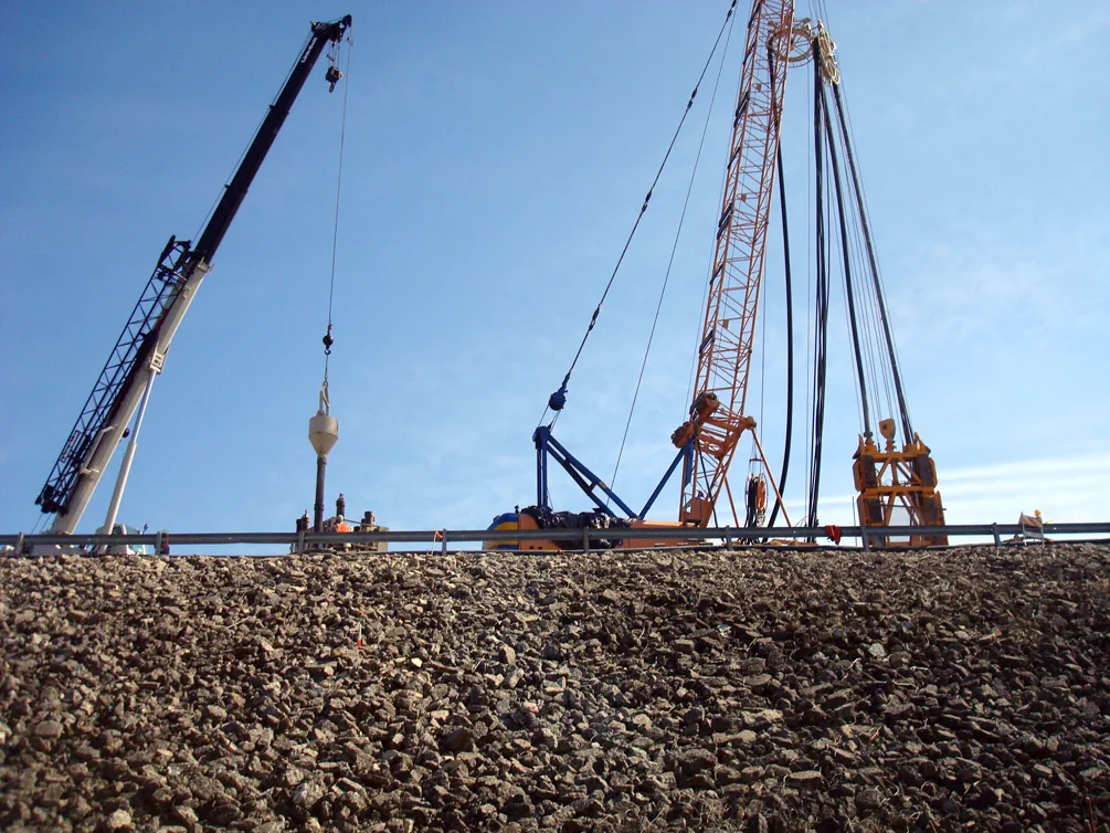 Distant view of crane and other construction equipment at the top of a rocky embankment; blue sky in background