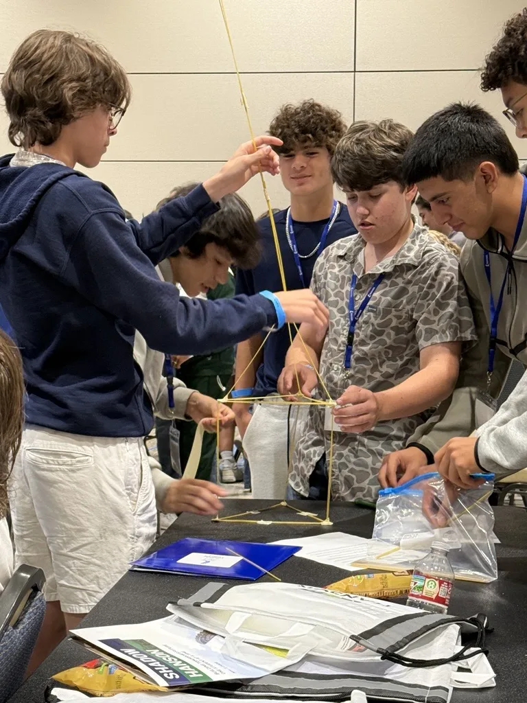 Six students standing around table, building structure with dry spaghetti noodles and marshmallows
