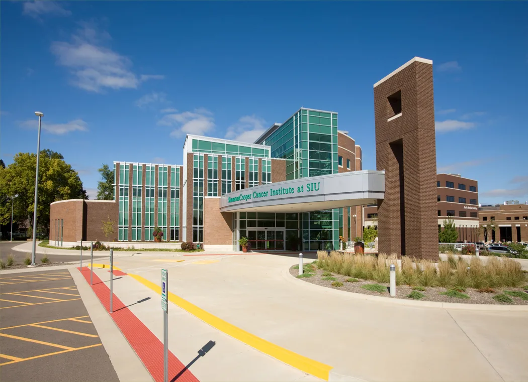 Exterior photo of medical center with sign on canopy reading, "SimmonsCooper Cancer Institute at SIU," green-tinted windows and brick facade, including brick tower supporting right side of canopy