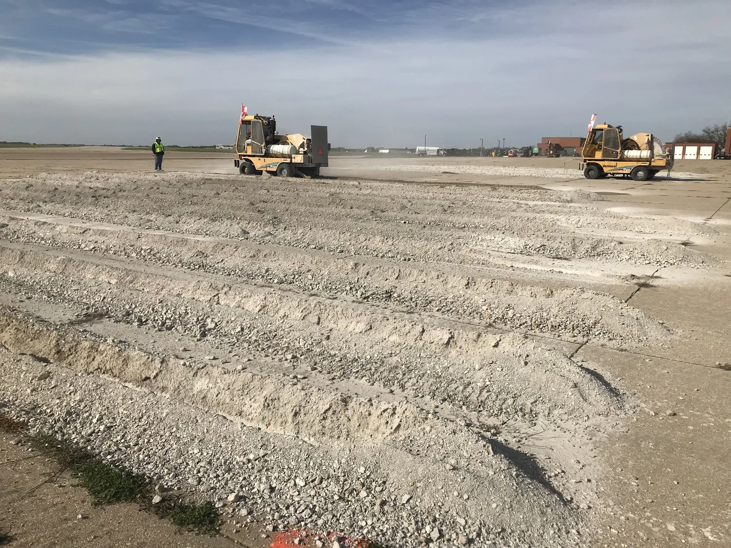 Rows of crushed concrete in foreground with two yellow construction vehicles in background along with a construction worker wearing a neon yellow safety vest and hard hat