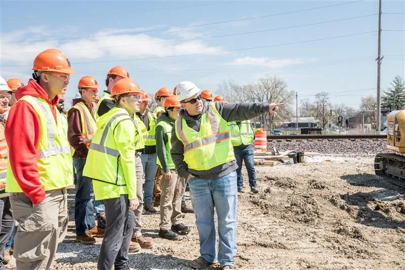 Group of construction workers in neon yellow safety vests and hard hats look toward right of screen, one pointing to something offscreen. Railroad track in background and part of yellow construction equipment at right