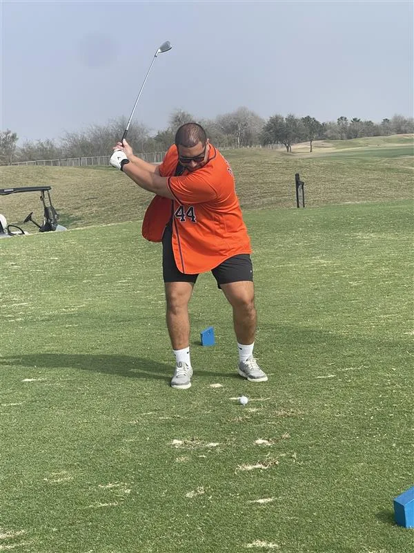 Man in orange baseball jersey and black shorts swinging golf club over ball with green grass in foreground and background