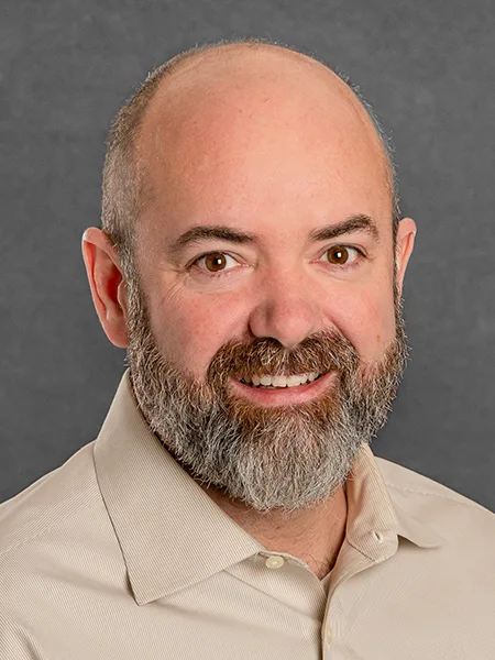 headshot of justin dewey wearing beige polo shirt on gray background