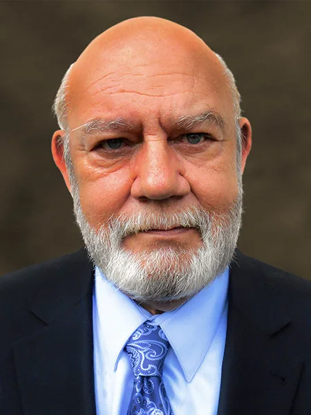 headshot of mike moon, wearing black suit jacket and light blue shirt with royal blue patterned tie on dark brown background