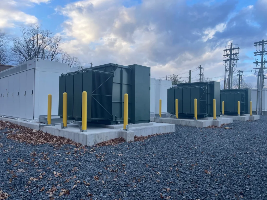 Battery yard energy storage system units lined up on concrete slabs with power lines in background and purple rocks in foreground