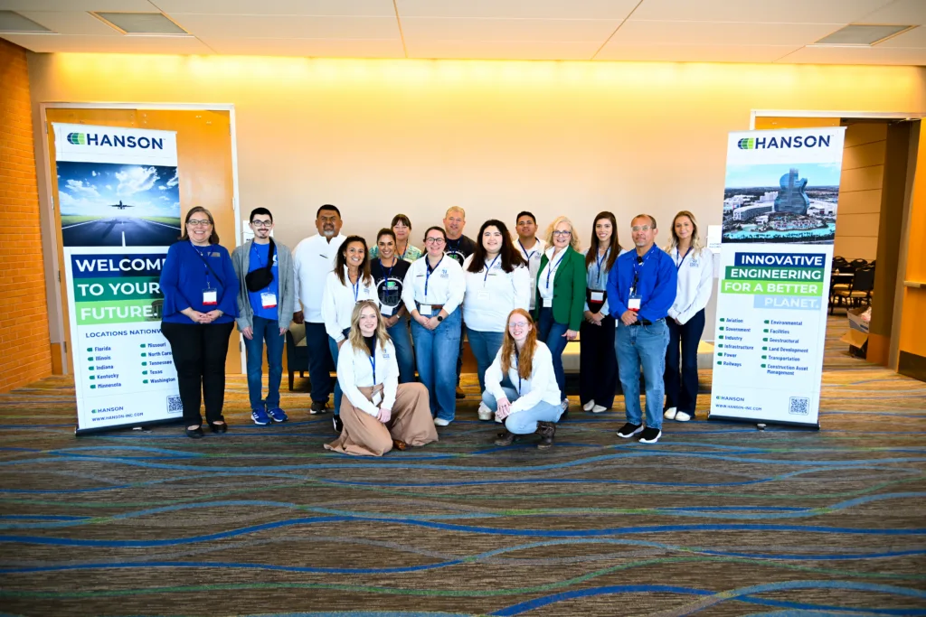 Group photo of people standing in front of yellow, top-lit wall between two tall banners with Hanson logo at top