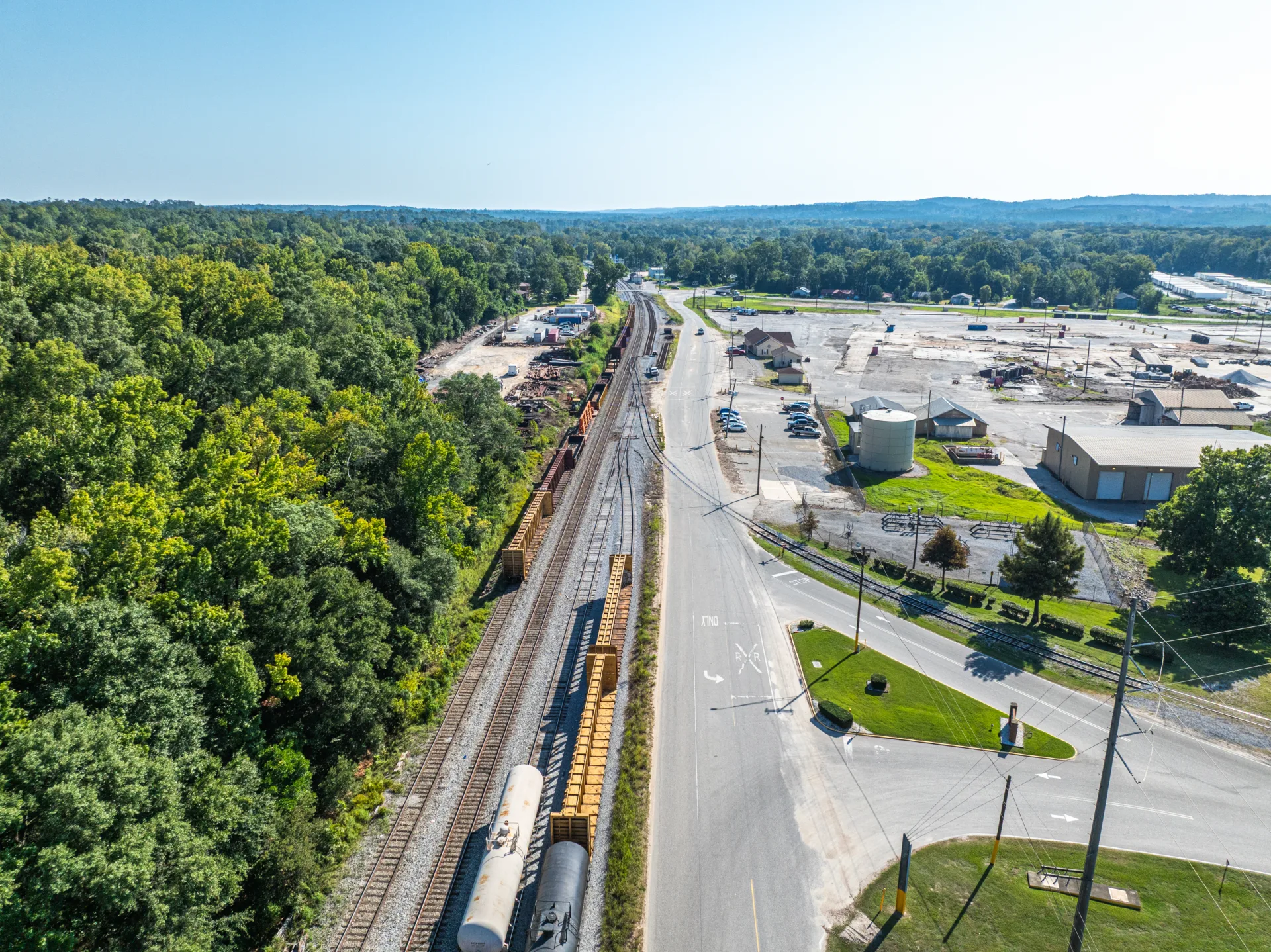 Aerial view of several sets of railroad tracks with new siding holding railroad cars; trees at left and industrial buildings at right; blue sky in background