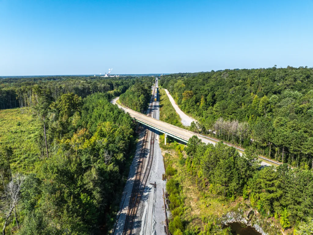 Aerial view of two sets of railroad tracks running under a bridge crossing over at an angle; trees on all sides and blue sky in background