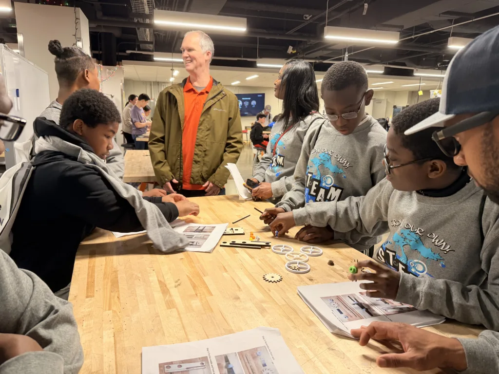 Photo of several students in gray sweatshirts sitting around a table, working on an experiment in a room with other tables and people in background