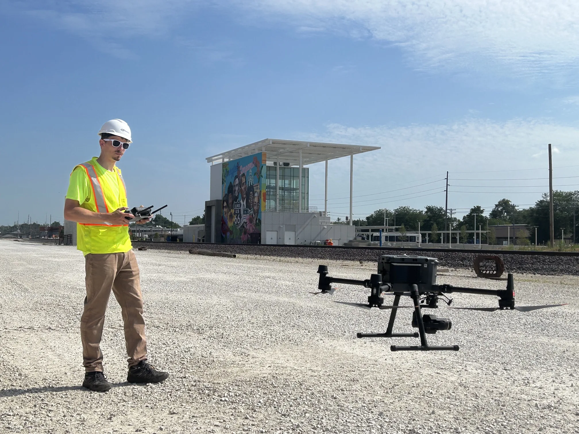 Man in tan pants and bright yellow safety vest and white hard hat holds controls to drone hovering about rocky ground to his right. In background is a set of railroad tracks and a building with a colorful mural
