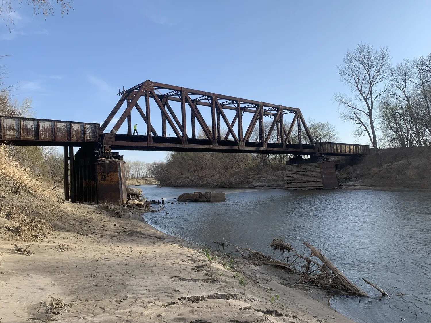 Brown truss bridge over river with dirt in foreground at left and trees on riverbank at right.