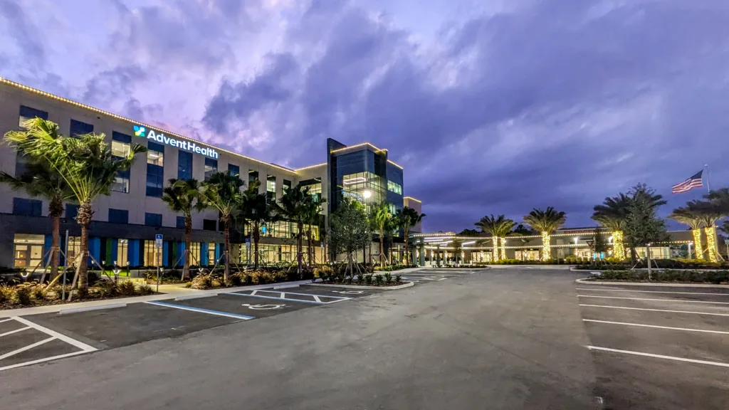 Exterior view of AdventHealth hospital at dusk with palm trees from left to right and American flag flying at right, empty parking lot in foreground