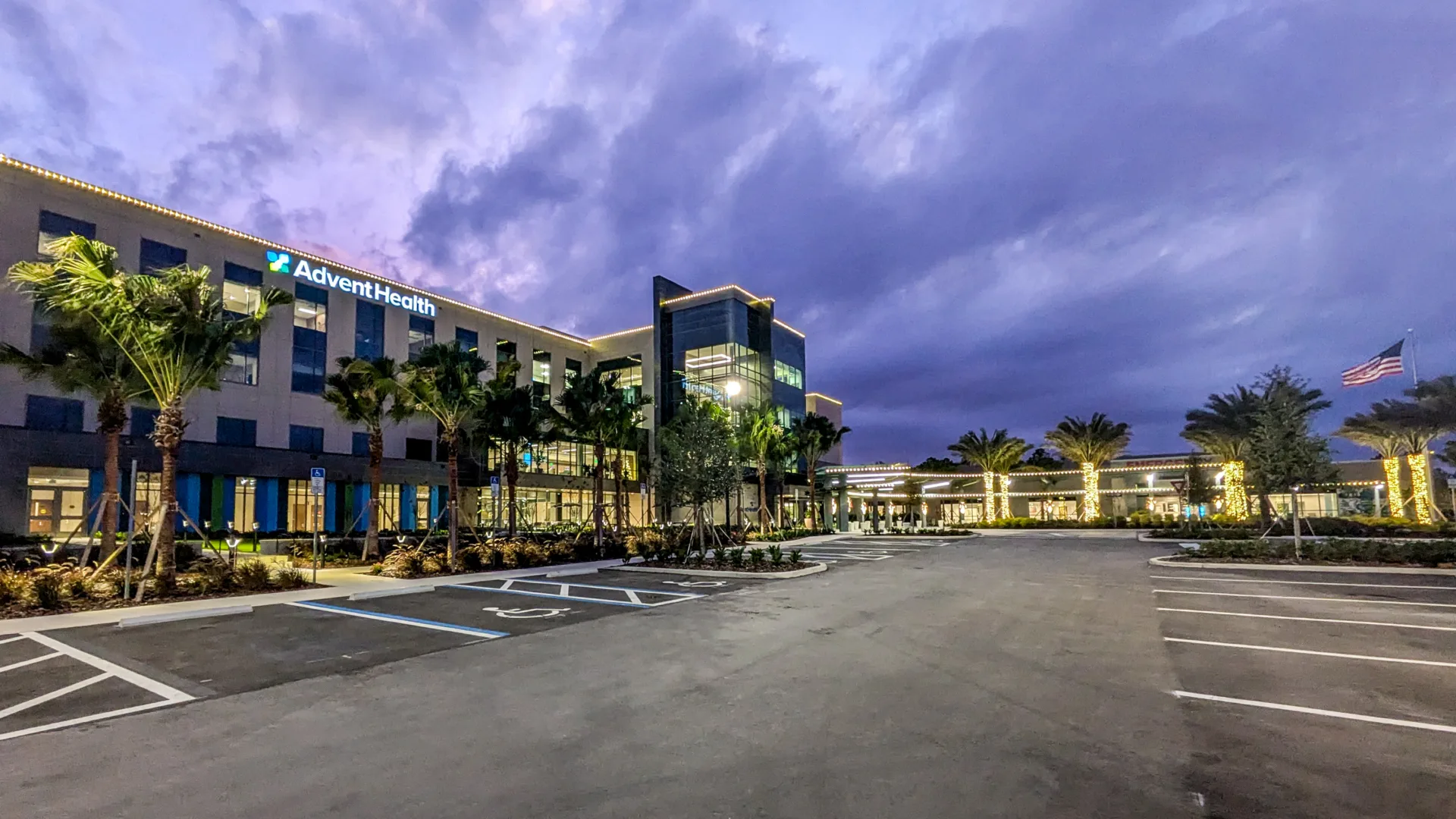 Exterior view of AdventHealth hospital at dusk with palm trees from left to right and American flag flying at right, empty parking lot in foreground