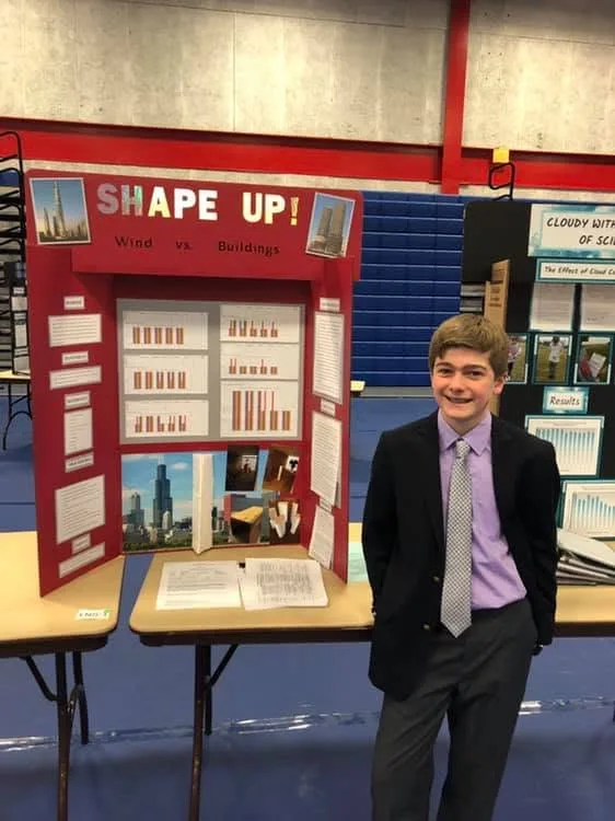 Boy wearing dark suit with purple button-down shirt and gray patterned tie stands in front of table next to exhibit board with title "Shape up!" at top, charts in middle, photos of skyscrapers at bottom and blurbs of text on right and left sides. Blue floor; blue bleachers and concrete wall with red trim in background