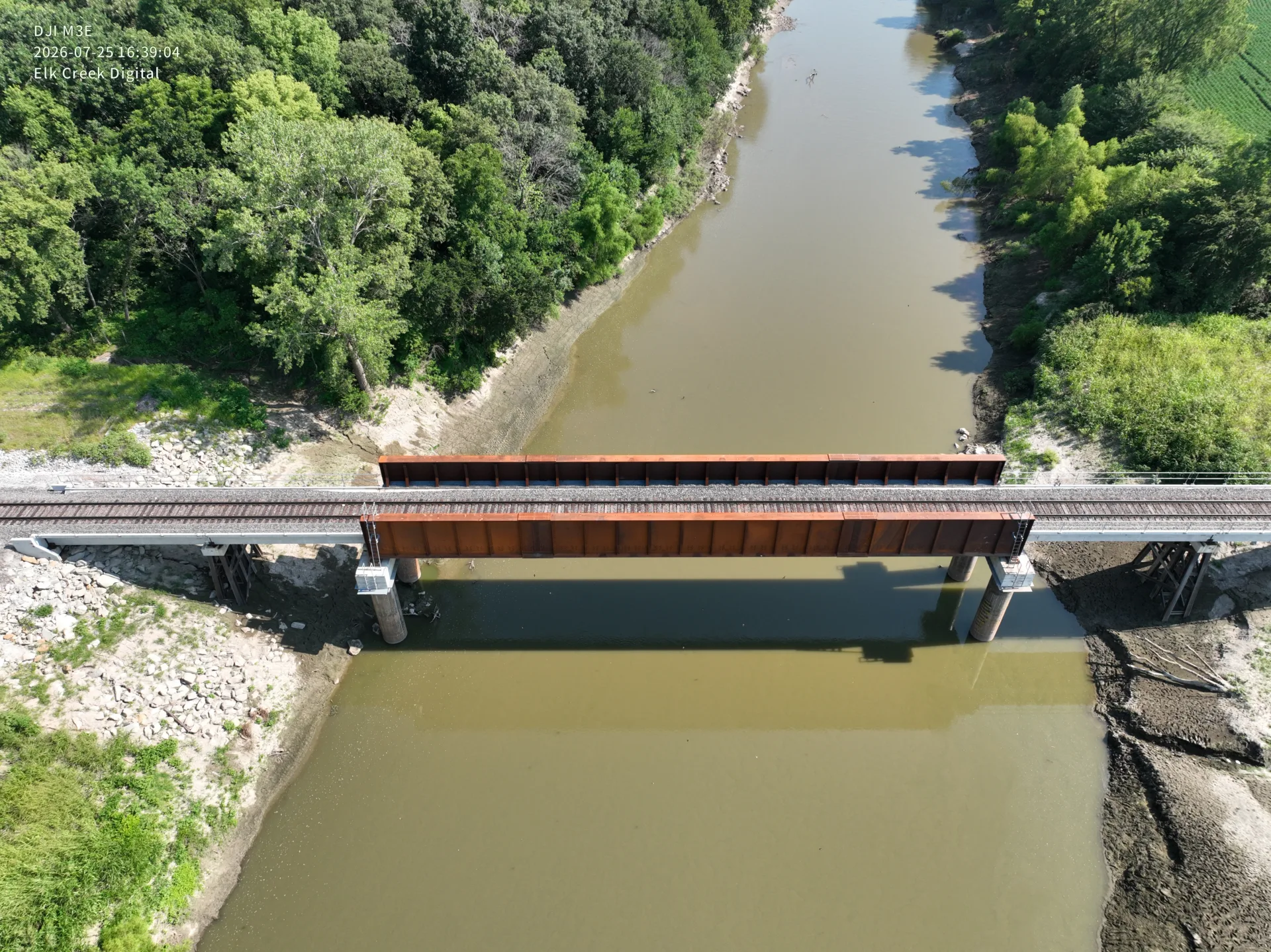 Aerial view of new railroad bridge, orange-brown in color, over river with green trees on either side.