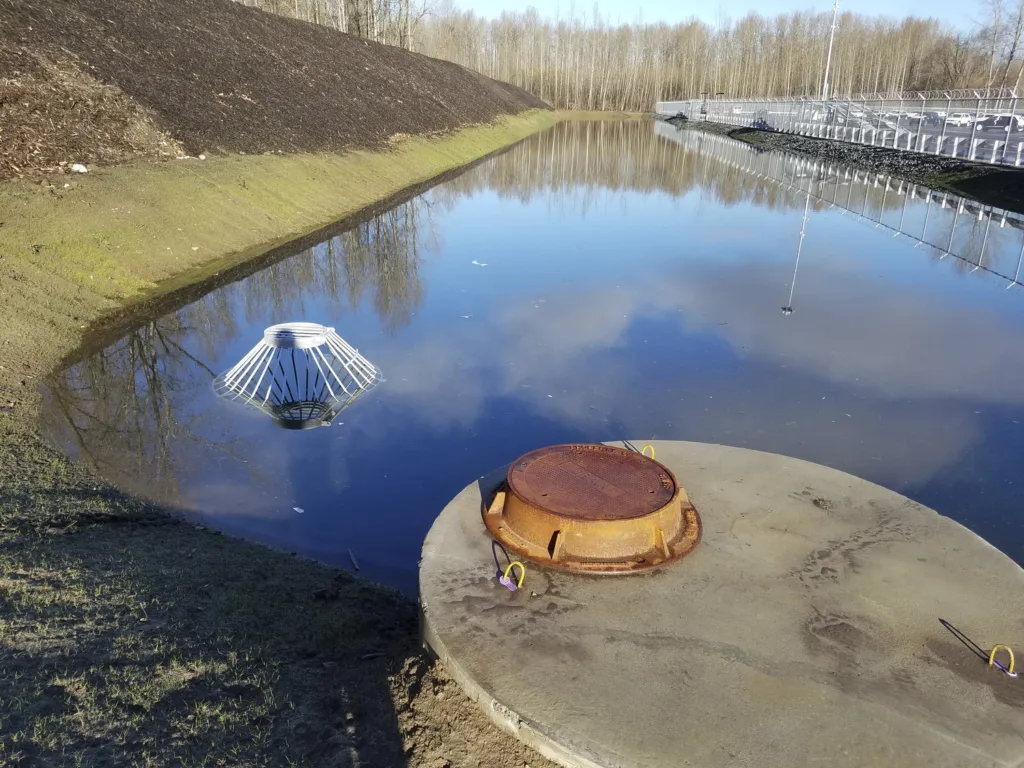 Photo of a retention pond with a covered manhole on a concrete slab in the foreground and rows of parked cars in a parking lot behind a fence to the right