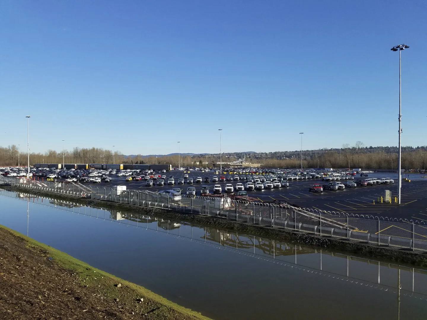 View of an auto yard with rows of parked cars and train cars in the background; retention pond in the foreground, separated from the parking lot by a fence