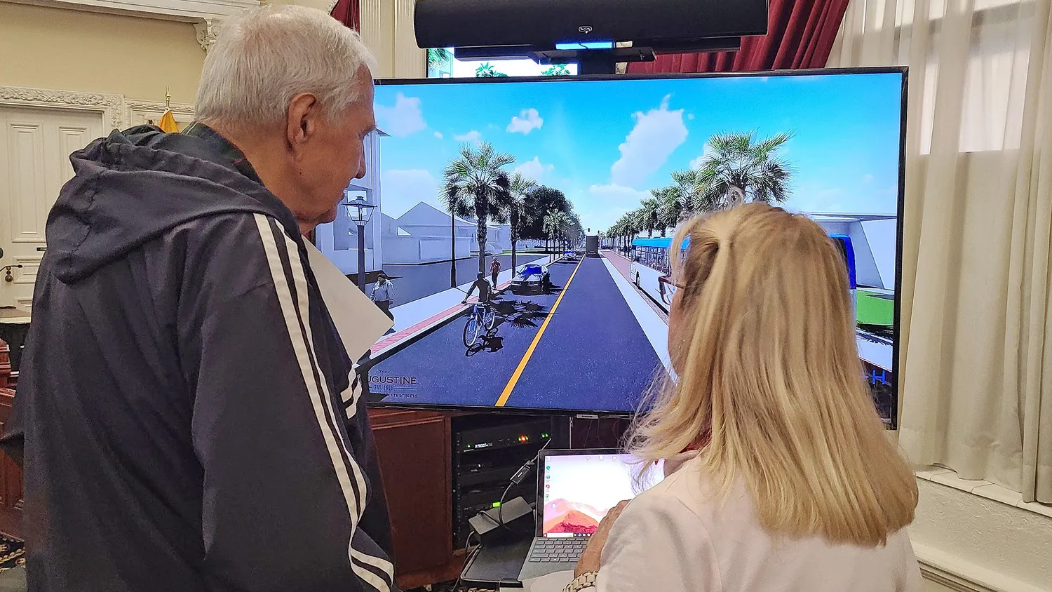 Backs of tall man with gray hair and shorter woman with long, blond hair and glasses as they observe a monitor with renderings of a street project with a cyclist, pedestrian and car on a street lined with palm trees