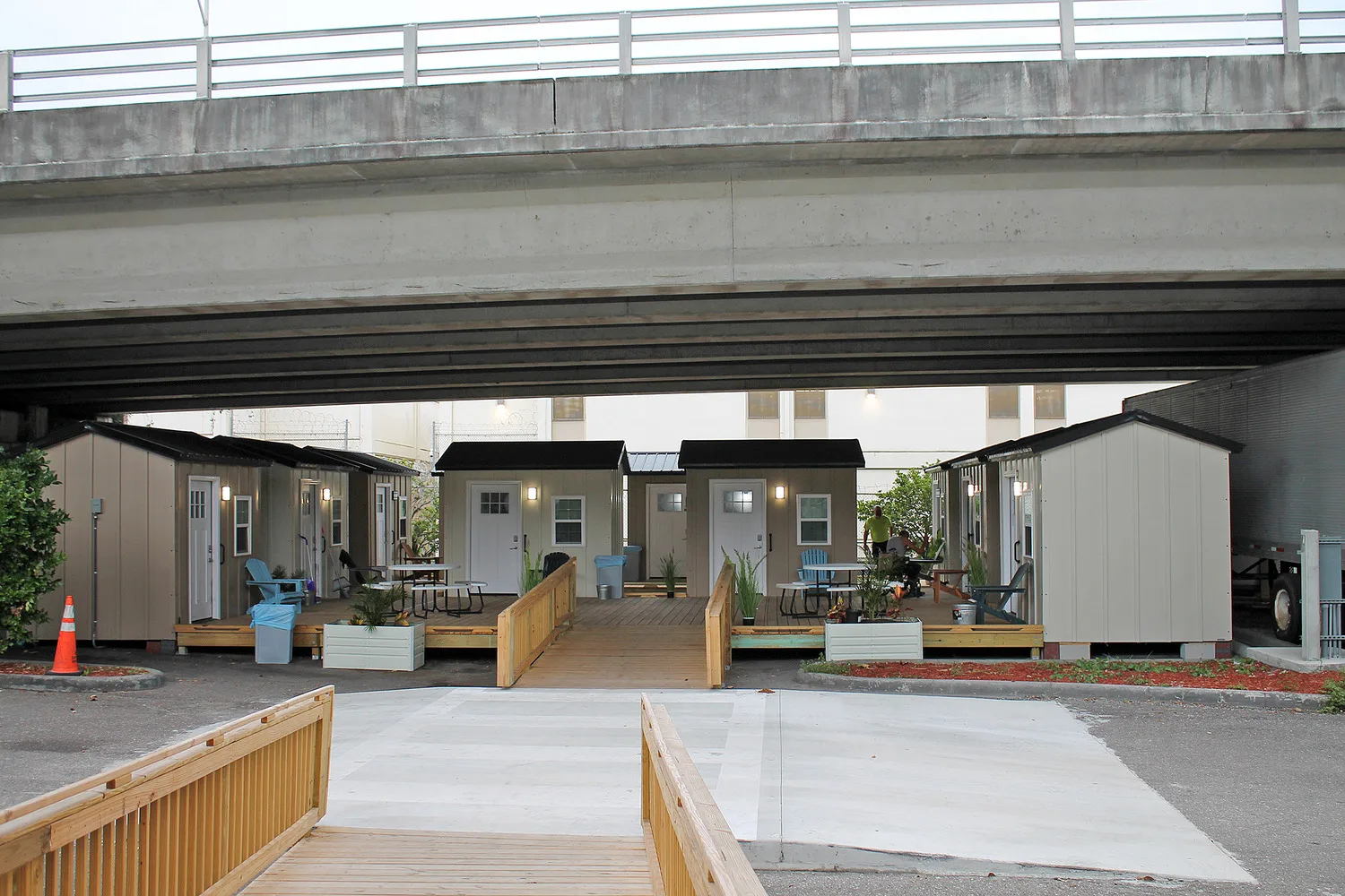 Photo of several tiny homes under an overpass with landscaping and a wooden ramp in foreground; small houses with white doors and white-framed windows lined up in background; white picnic table in front of one house