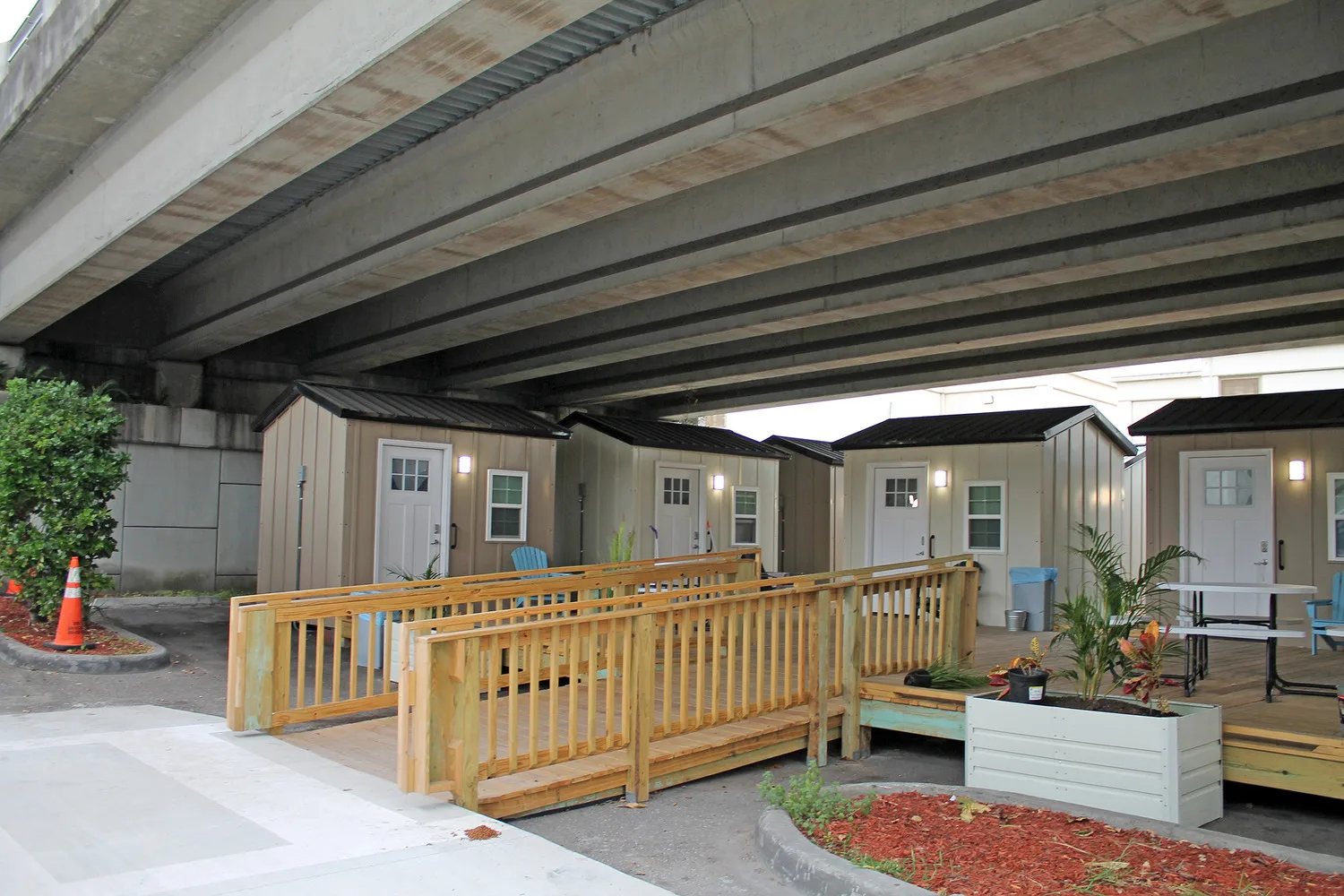 Photo of several tiny homes under an overpass with landscaping and a wooden ramp in foreground; small houses with white doors and white-framed windows lined up in background; white picnic table in front of one house
